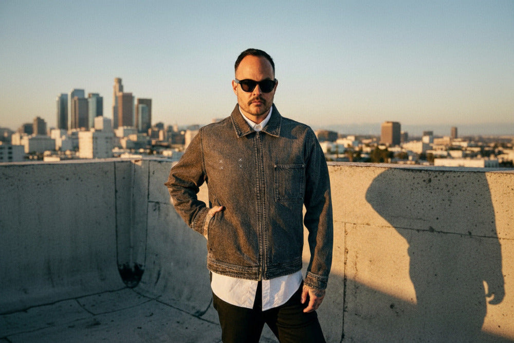 Man wearing a denim jacket and sunglasses on a rooftop with a cityscape in the background